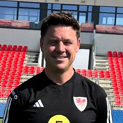 Mark smiling facing the camera in a stadium with black football kit on.